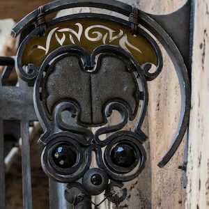 Puede incluir: Un letrero de metal negro con una cara estilizada y la palabra "Vongola" escrita en oro sobre un fondo amarillo. El letrero está sujeto a una pared con un soporte de metal.