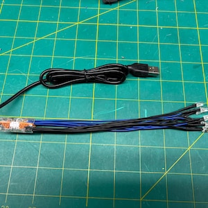 May include: A black USB cable connected to a bundle of black and blue wires with silver connectors. The wires are attached to a clear plastic component with orange and white details. The items are on a green cutting mat.
