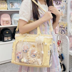 Puede incluir: Un bolso tote amarillo claro con un panel frontal transparente, que muestra pines decorativos y un adorno esponjoso. El bolso tiene una correa para el hombro y un ribete de encaje. Se ven otros bolsos en estantes en el fondo.