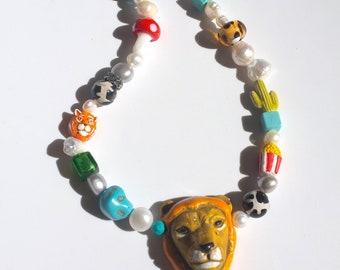May include: A colorful beaded necklace with a lion head charm. The necklace features a variety of beads, including white pearls, turquoise, green, red, orange, and black. The lion head charm is made of orange and brown ceramic.