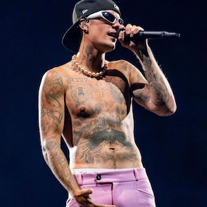 Könnte beinhalten: Ein Mann mit einer schwarzen Baseballkappe, einer weißen Sonnenbrille und einer Perlenkette, mit zahlreichen Tattoos auf Brust und Armen, singt in ein Mikrofon. Er trägt eine rosa Hose und weiße Unterwäsche.