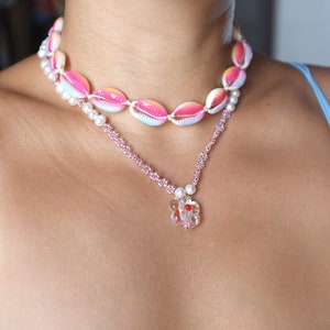 May include: Two necklaces, one with pink and white seashells and the other with pink beads and a clear flower charm with red accents.