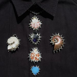 May include: A collection of unique shell pendants with silver-toned, spiky metalwork. The pendants feature various shell shapes and colors, including white, pink, brown, and blue, set against a black background.