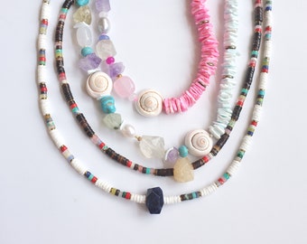 May include: A layered necklace with multiple strands of beads, including white, pink, and brown beads. The necklace features various shapes and colours, including turquoise, pink, and white shells, as well as amethyst and quartz crystals.