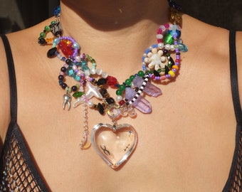 May include: A colorful beaded necklace with a large glass heart pendant. The necklace features a variety of beads, including pearls, crystals, and glass beads. The heart pendant is clear and has a delicate design.