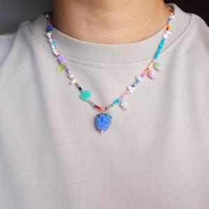 Puede incluir: Un collar de cuentas de colores con un dije de baya azul de vidrio. El collar presenta una variedad de cuentas, incluyendo perlas, corazones y flores.