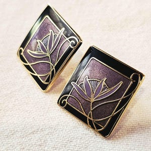 May include: A pair of gold-toned metal earrings featuring a diamond-shaped design with a black border and a textured purple center. Each earring displays a stylized floral motif in light purple and gold lines, creating a geometric aesthetic.