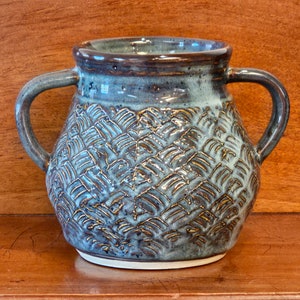 May include: A handcrafted ceramic vase with two handles. The vase features a textured, woven pattern in shades of blue and brown. The interior and rim are a darker blue, and the base is white.