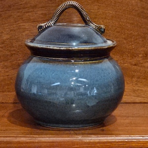May include: A blue ceramic jar with a lid and a woven handle. The jar has a rounded shape and a glossy finish. The lid is dark blue with a gold trim. The handle is a twisted, rope-like design.
