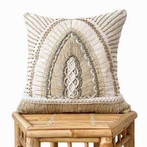 Puede incluir: Una almohada de color beige y blanco con un dise&ntilde;o de arco tejido y ribete de flecos. La almohada est&aacute; sentada en un taburete de madera marr&oacute;n claro.