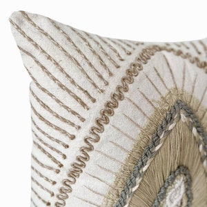 Puede incluir: Una almohada blanca con un dise&ntilde;o bordado en beige y marr&oacute;n. El dise&ntilde;o presenta un patr&oacute;n geom&eacute;trico con un c&iacute;rculo central.