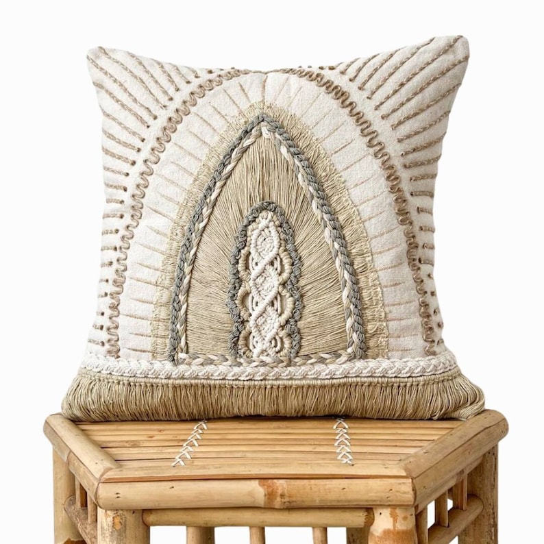 Puede incluir: Una almohada de color beige y blanco con un dise&ntilde;o de arco tejido y ribete de flecos. La almohada est&aacute; sentada en un taburete de madera marr&oacute;n claro.