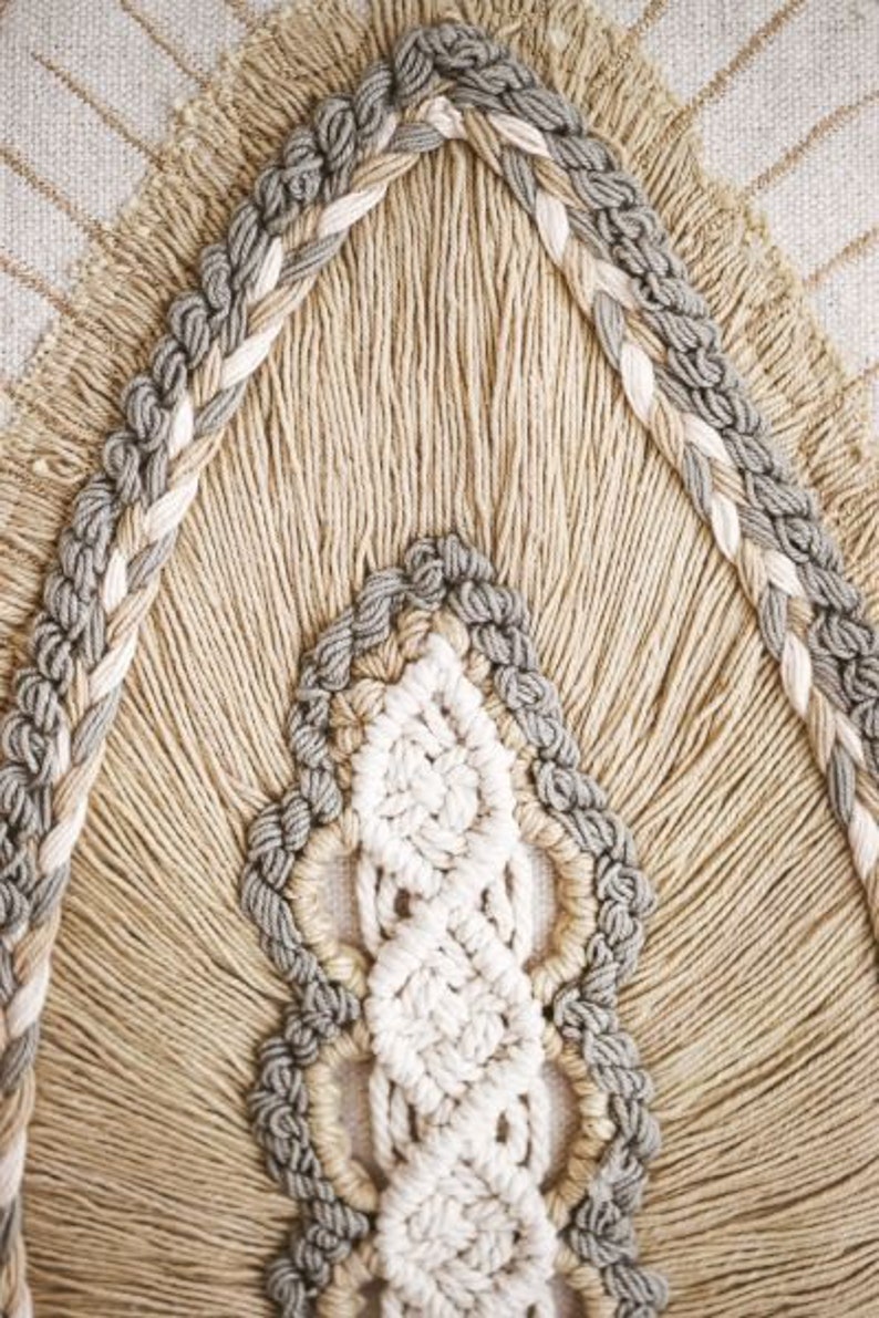 Puede incluir: Primer plano de un tapiz de macram&eacute; beige y gris con un patr&oacute;n tejido. El macram&eacute; est&aacute; hecho con fibras naturales gruesas y tiene un borde con flecos.