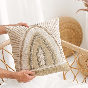 Puede incluir: Una almohada de color beige y marr&oacute;n con un dise&ntilde;o tejido y ribete de flecos. La almohada tiene un patr&oacute;n geom&eacute;trico con una gran forma de arco en el centro.