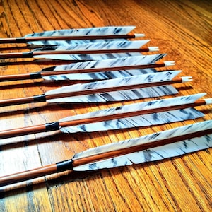 May include: A collection of wooden arrows with white and gray fletching, arranged on a wooden surface. The arrows have brown shafts, black bindings, and white tips. The fletching features a marbled pattern.