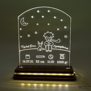 Può includere: Luce notturna illuminata con un disegno inciso de Il Piccolo Principe, stelle e una luna crescente. La base mostra la data 16.07.21, una lunghezza di 52 cm, l'ora 11:15 e un peso di 3200 g. Realizzata in legno e acrilico.