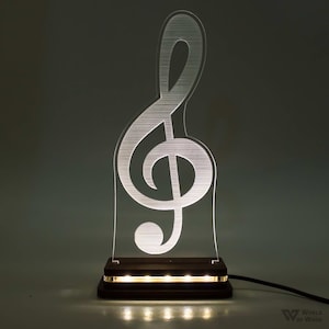 Può includere: Lampada a forma di chiave di violino musicale illuminata a LED. La chiave di violino è in acrilico trasparente e poggia su una base in legno scuro. Il cavo di alimentazione è nero. L'altezza della lampada è di circa 25 cm.