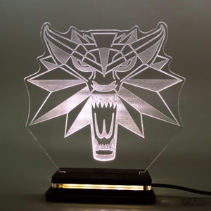 Può includere: Una lampada in acrilico trasparente con un design geometrico a testa di lupo, illuminata da una luce calda. La lampada poggia su una base in legno scuro di forma rettangolare. Il design è ispirato alla serie The Witcher.