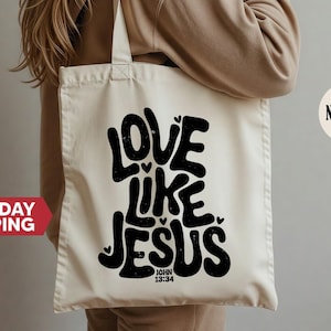 May include: Cream-colored tote bag with the words "LOVE LIKE JESUS" in bold black lettering. The text includes small heart shapes and the Bible verse John 13:34. The bag has a simple design and is carried over the shoulder.