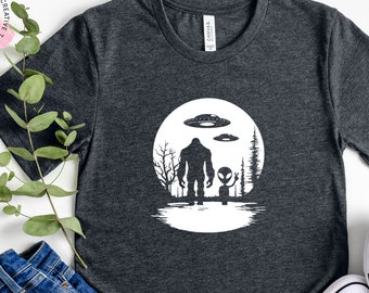 Könnte beinhalten: Ein dunkelgraues T-Shirt mit einem weißen Aufdruck von Bigfoot und einem Außerirdischen, die vor einem Vollmond mit fliegenden Untertassen stehen.