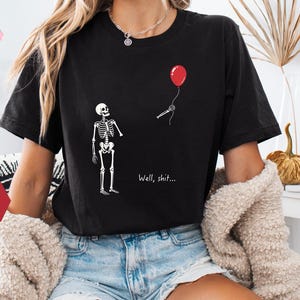 May include: A black short-sleeve t-shirt featuring a white graphic of a skeleton looking up at a red balloon that is floating away. The text "Well, shit..." is printed in white below the balloon. The shirt has a crew neckline and a relaxed fit.