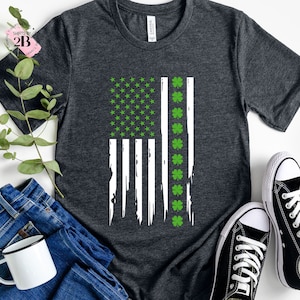 May include: A dark heather grey t-shirt featuring a distressed American flag design in white and green. The flag has shamrocks replacing the stars on the blue field.