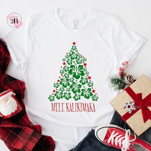 May include: White t-shirt with a green Christmas tree made of red and green hibiscus flowers. The text "Mele Kalikimaka" is printed below the tree.