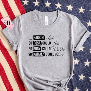 May include: Gray t-shirt with black text that reads "... HARRIET Led SO ROSA COULD Sit SO RUBY COULD Walk SO KAMALA COULD Run" on a red, white, and blue American flag background.