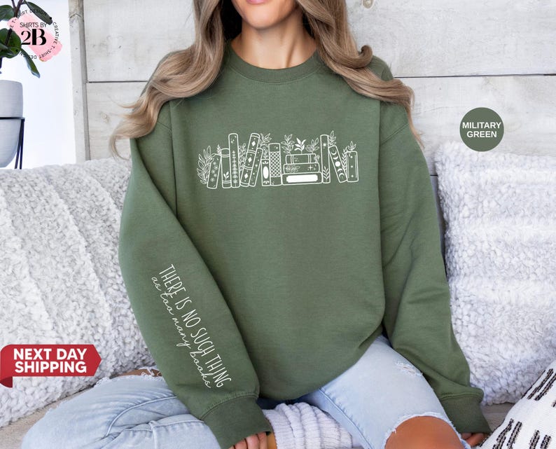 May include: Military green sweatshirt with a white graphic of stacked books and foliage. The sleeve features the text "THERE IS NO SUCH THING as too many books" in white. The sweatshirt is a comfortable, casual style.