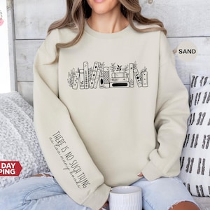 May include: A sand-coloured sweatshirt with a black graphic of books and foliage. The left sleeve has the text "THERE IS NO SUCH THING as too many books." The sweatshirt is a neutral colour and has a crew neck.