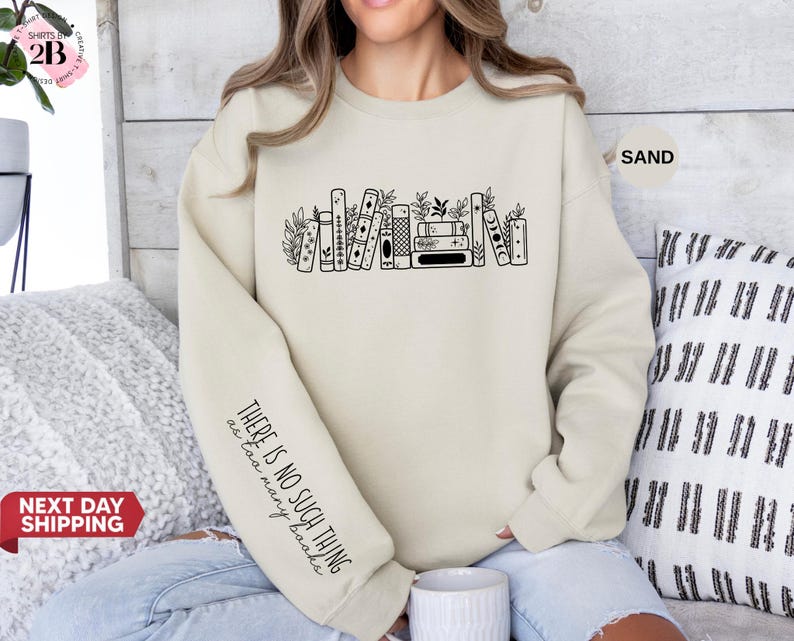 May include: A sand-coloured sweatshirt with a black graphic of books and foliage. The left sleeve has the text "THERE IS NO SUCH THING as too many books." The sweatshirt is a neutral colour and has a crew neck.
