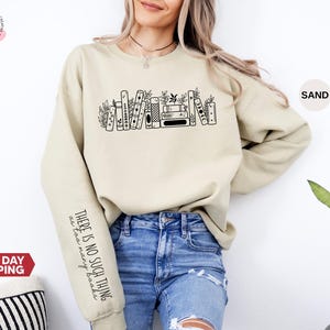 May include: A sand-coloured sweatshirt featuring a black bookshelf graphic and the text "THERE IS NO SUCH THING as too many books" on the sleeve. The sweatshirt is made of a soft material and is suitable for casual wear.