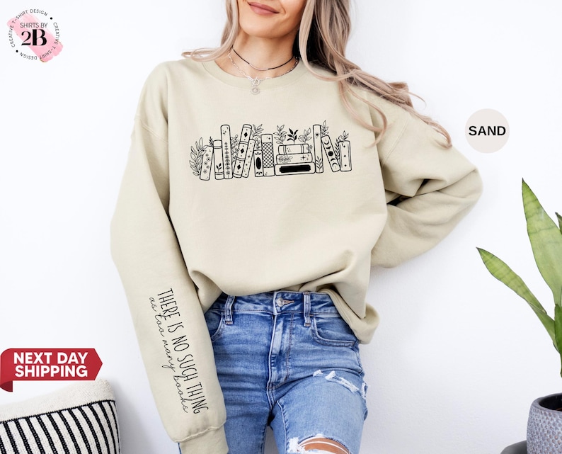 May include: A sand-coloured sweatshirt featuring a black bookshelf graphic and the text "THERE IS NO SUCH THING as too many books" on the sleeve. The sweatshirt is made of a soft material and is suitable for casual wear.
