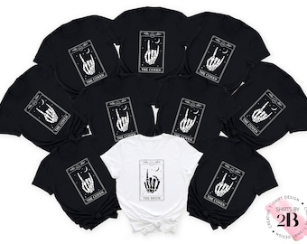 May include: A set of black t-shirts featuring a white graphic of a skeleton hand making a rock and roll sign. The shirts have the text "The Coven" printed on them. One white t-shirt has the text "The Bride" printed on it.