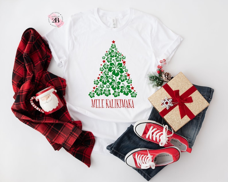 May include: White t-shirt with a green Christmas tree made of red and green hibiscus flowers. The text "Mele Kalikimaka" is printed below the tree.