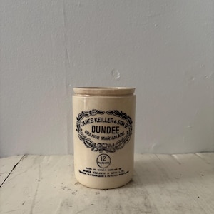 May include: A cylindrical cream-coloured ceramic jar featuring black printed text that reads James Keiller & Son Dundee Orange Marmalade inside a decorative wreath. Below the wreath, a circle contains the text 12 FLUID OZS.