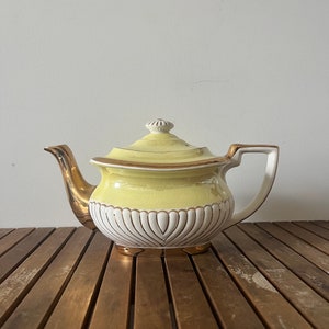 Puede incluir: Una tetera amarilla con ribetes dorados y detalles blancos. La tetera tiene un cuerpo redondeado y una tapa con un pomo en la parte superior. La tetera está sobre una superficie de madera.