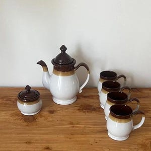 May include: A ceramic tea set comprising a teapot, sugar bowl, and four mugs. The set has a white base with brown and tan detailing. The teapot features a curved spout and handle, and the mugs are rounded.