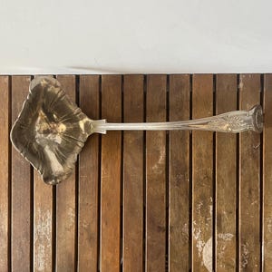 Peut inclure: Une louche argentée avec un manche décoratif et un large bol cannelé. La louche repose sur une surface en bois avec des planches verticales. La poignée présente un motif orné.