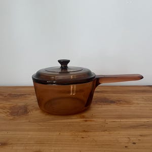 Puede incluir: Una cacerola vintage de vidrio color ámbar con tapa y asa. El vidrio transparente permite ver el contenido. El asa y el pomo de la tapa también son de color ámbar. Esta batería de cocina es adecuada para usar en la estufa. Aproximadamente 15 cm de diámetro.