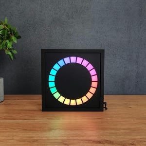 Op de afbeelding: Een zwarte vierkante digitale klok met een cirkelvormige weergave van kleurrijke lichten. De klok staat op een houten oppervlak, met een plant in een pot op de achtergrond. De lichten tonen een regenboog van kleuren.