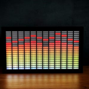 May include: A black LED sound visualizer with a colourful display. Vertical bars of light change from red at the top to yellow and blue at the bottom. The device sits on a wooden surface. The visualizer is designed to react to sound, creating a dynamic display.
