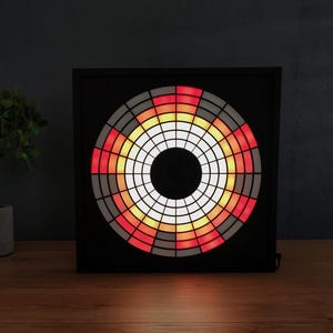 Op de afbeelding: Een zwart vierkant LED-licht met een cirkelvormig, gesegmenteerd ontwerp. De segmenten zijn gekleurd in een verloop van rood, oranje, geel en wit. Het licht staat op een houten oppervlak, met een plant in een pot links.
