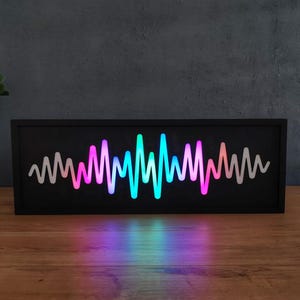 Peut inclure: Une œuvre d'art sonore rectangulaire noire avec un motif d'égaliseur lumineux et coloré. Le motif des ondes passe du rose au bleu et au vert. L'œuvre est présentée sur une surface en bois.