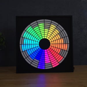 Op de afbeelding: Een vierkant zwart wanddisplay met een cirkelvormig lichtpaneel met een radiaal rasterpatroon. Het raster bestaat uit meerdere segmenten die oplichten in een kleurenspectrum waaronder blauw, groen, geel, oranje, rood en paars.