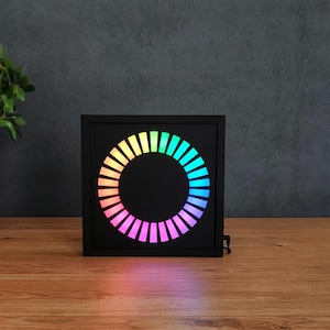 May include: A black square LED clock with a circular rainbow light display. The clock sits on a wooden surface, with a potted plant in the background. The light display transitions through various colors.