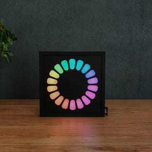 May include: A square black frame containing a circular display of twenty-four illuminated segments. The segments transition in colour from cyan and blue at the top to purple, magenta, and orange at the bottom, creating a vibrant gradient effect.