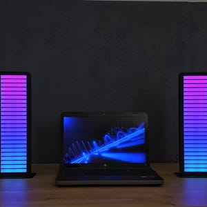 Op de afbeelding: Twee verticale rechthoekige lichtbalken met een verloop van paarse naar blauwe led-segmenten, geplaatst aan weerszijden van een zwarte laptop. De lichtbalken hebben een rasterachtig display van verlichte horizontale lijnen in een donkere omgeving.