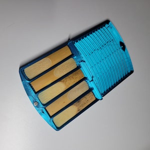 May include: A blue case with four wooden clarinet reeds inside. The case has a textured blue surface and a magnetic closure.