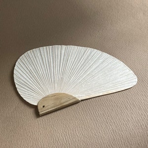 Può includere: Un ventaglio di carta bianca con manico in legno. Il ventaglio ha un design a pieghe, che si irradia verso l'esterno dal manico. Il ventaglio è di colore crema chiaro ed è aperto, mostrando la sua forma completa.